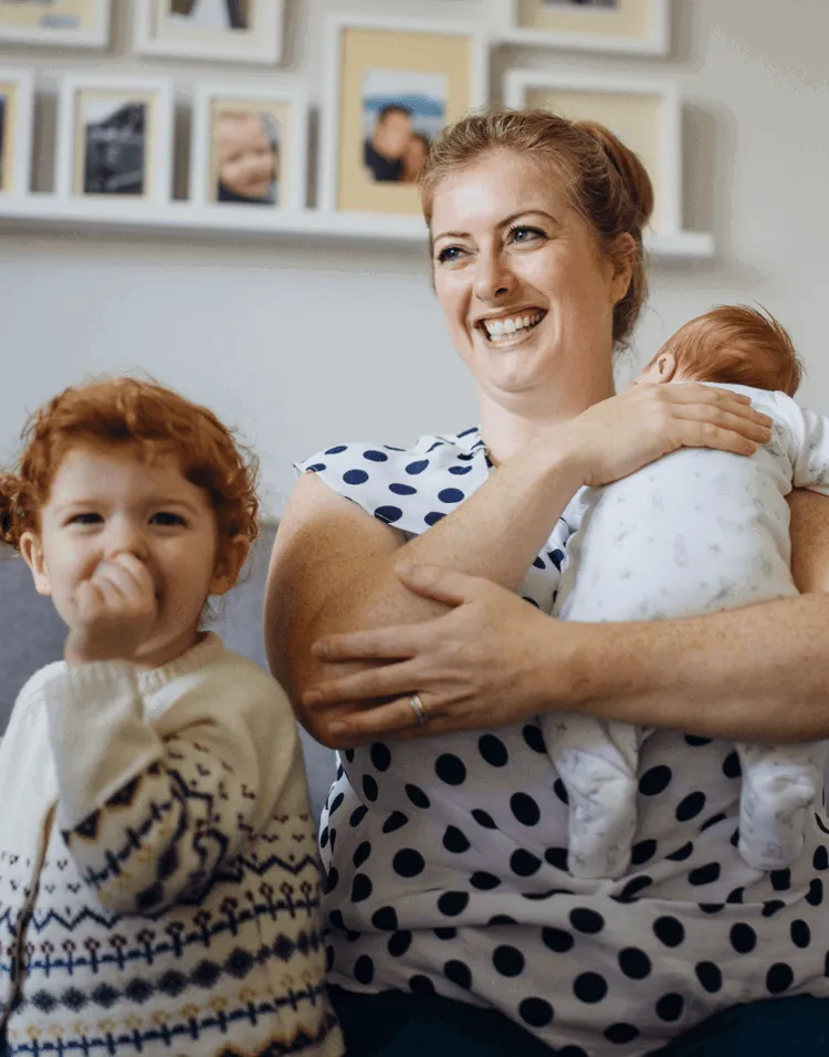 A smiling mother at home holding her baby, with her young child beside her.